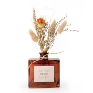 May include: A square amber glass diffuser bottle with dried floral arrangement. The bottle has a label that reads "Amber Vanilla, Hand Poured, Botanical, Reed Bundles, 7 fl oz (207 ml)". The arrangement includes wheat, orange flowers, and fluffy beige accents.
