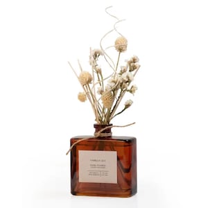 May include: A brown glass diffuser bottle with dried flowers and reeds. The bottle has a rectangular shape and a label that reads "Vanilla Lily Hand Poured Luxury Diffuser". The flowers are beige and white, and tied with twine.