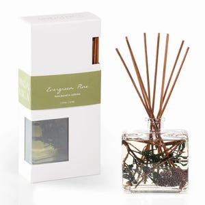 May include: A clear square glass reed diffuser bottle filled with liquid, pine needles, and a small pinecone, featuring several brown wooden reeds. Beside it stands a white rectangular box labeled Evergreen Pine, 7.5 fl oz, hand poured in California.