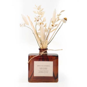 May include: A square amber glass reed diffuser bottle with a beige label reading WHITE TEA & PAMPAS HAND POURED LUXURY DIFFUSER BOTANICAL REED DIFFUSER 7 FL OZ (207 ML). The bottle is tied with twine and contains dried white flowers and pampas grass.