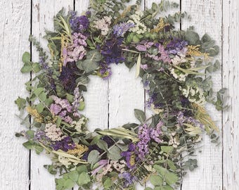 May include: A circular wreath made from dried flowers and foliage. The wreath displays a mix of colours, including green, purple, and yellow. The wreath is composed of eucalyptus leaves, lavender, and other dried flowers.