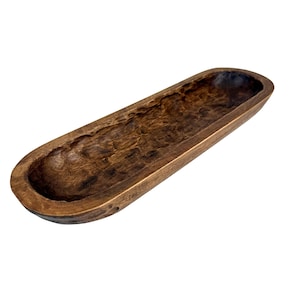May include: A long, dark brown wooden bowl with a textured interior. The bowl has an oval shape and is made of a single piece of wood. The exterior is smooth, while the interior has a carved, rustic appearance. This decorative bowl is suitable for various uses.