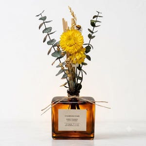 May include: A square amber glass reed diffuser bottle with a white label reading GOLDEN BLOOMS HAND POURED LUXURY DIFFUSERS BOTANICAL REED BUNDLES 7 FL OZ (207 ML). The bottle is tied with twine and contains yellow flowers and eucalyptus.