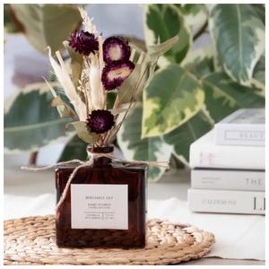 May include: A square amber glass reed diffuser bottle with a white label reading Bergamot Lily, Hand Poured, 7 fl oz (207 ml). The bottle is tied with twine and contains dried purple flowers, white pampas grass, and wooden reeds.