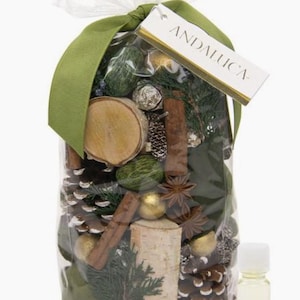 May include: A clear cellophane bag filled with potpourri, including pine cones, cinnamon sticks, and wooden accents. The bag is tied with a green ribbon and a tag that reads "ANDALUCA". A small bottle of essential oil is next to the bag.