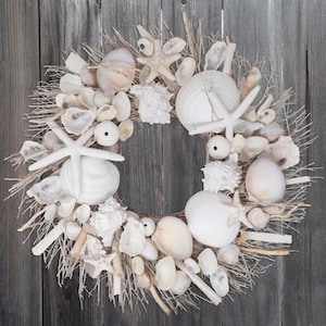 May include: A white seashell wreath with a variety of seashells, starfish, and driftwood. The wreath is made of natural materials and has a rustic, coastal feel.