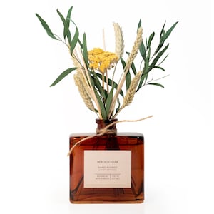 May include: A square amber glass reed diffuser bottle with a beige label reading NEROLI CEDAR HAND POURED LUXURY DIFFUSERS BOTANICAL REED BUNDLES 7 FL OZ (207 ML). A twine bow is tied around the neck, holding dried yellow flowers and green foliage.