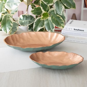 May include: Two decorative serving bowls with a copper-colored interior and a teal-colored exterior. The bowls are oval shaped and have a wavy edge.