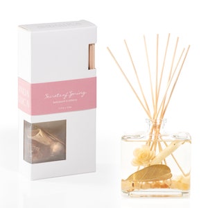 May include: A white box with a pink label that reads "Secrets of Spring" hand poured in California. The box contains a clear glass bottle with a gold leaf, white flower, and wooden sticks. The bottle is filled with a clear liquid.