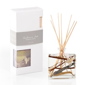 May include: A white box with a gray label that reads "Cashmere Oak" hand poured in California. The box contains a clear glass bottle with wooden sticks and a floral scent. The bottle is filled with a clear liquid and has a floral design.