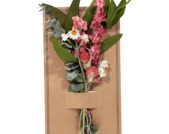 May include: A bouquet of dried flowers in shades of pink and white, arranged in a brown paper card with a small brown paper tag. The flowers include pink statice, white daisies, and green eucalyptus.