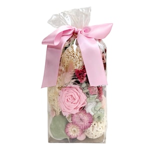 May include: A clear cellophane bag filled with dried flowers and greenery, tied with a light pink satin ribbon. The arrangement includes a large pink rose, various dried flowers, and green leaves. The bag is sealed at the top.