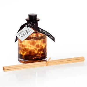 May include: A decorative glass diffuser bottle with a brown spotted pattern, black cap, and a black ribbon with the word "ANDALUCA." A tag reads "Cassis Vanilla." Several wooden reeds are placed in front of the bottle.