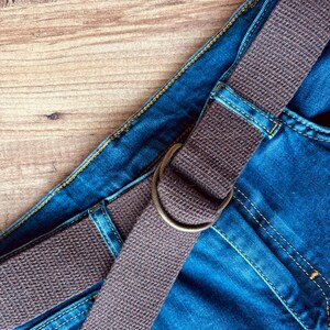 May include: A close-up of a brown canvas belt with a metal buckle, on top of a pair of blue denim jeans.