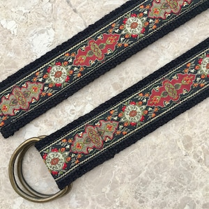 May include: A black woven belt with a gold and red patterned trim. The belt has a double-ring buckle.