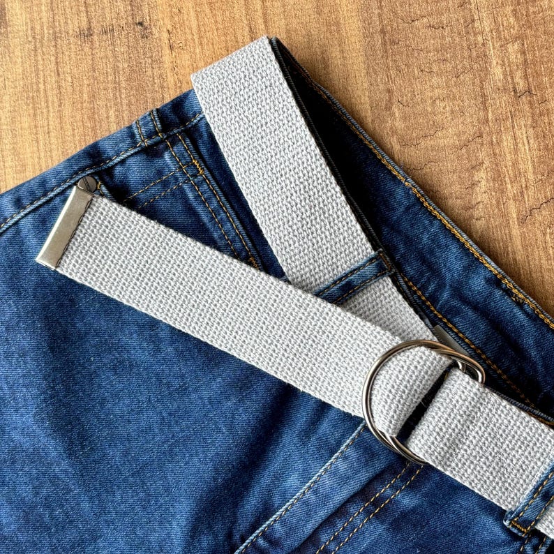 May include: A grey canvas belt with a silver buckle on a pair of blue denim jeans.