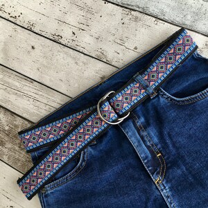 May include: A blue denim jean with a black and multi-colored patterned belt. The belt has a silver buckle.