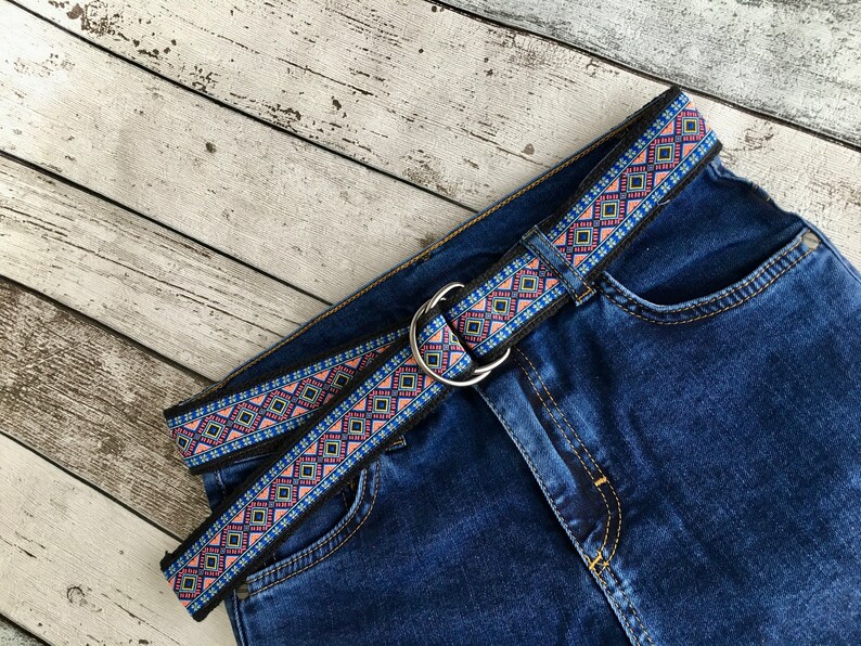 May include: A blue denim jean with a black and multi-colored patterned belt. The belt has a silver buckle.