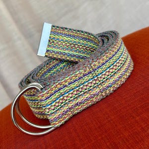 May include: A colorful woven belt with a silver double-ring buckle. The belt features a mix of colors including yellow, blue, green, and orange. The belt is rolled up on a red surface.