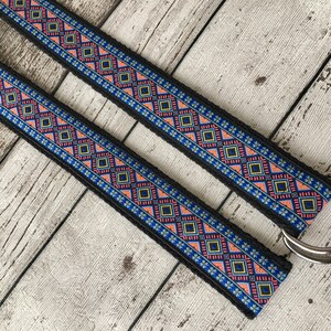 May include: A black woven belt with a colorful geometric pattern in shades of blue, pink, and yellow. The belt has a silver metal D-ring.