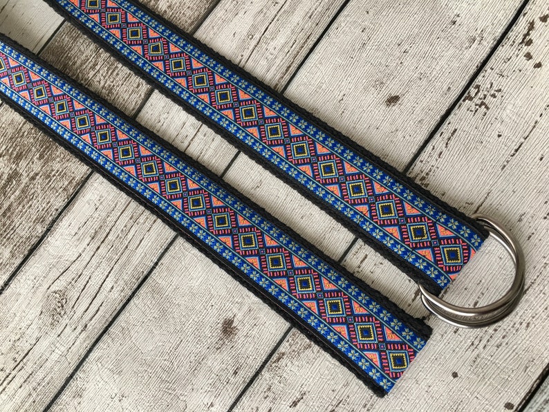 May include: A black woven belt with a colorful geometric pattern in shades of blue, pink, and yellow. The belt has a silver metal D-ring.
