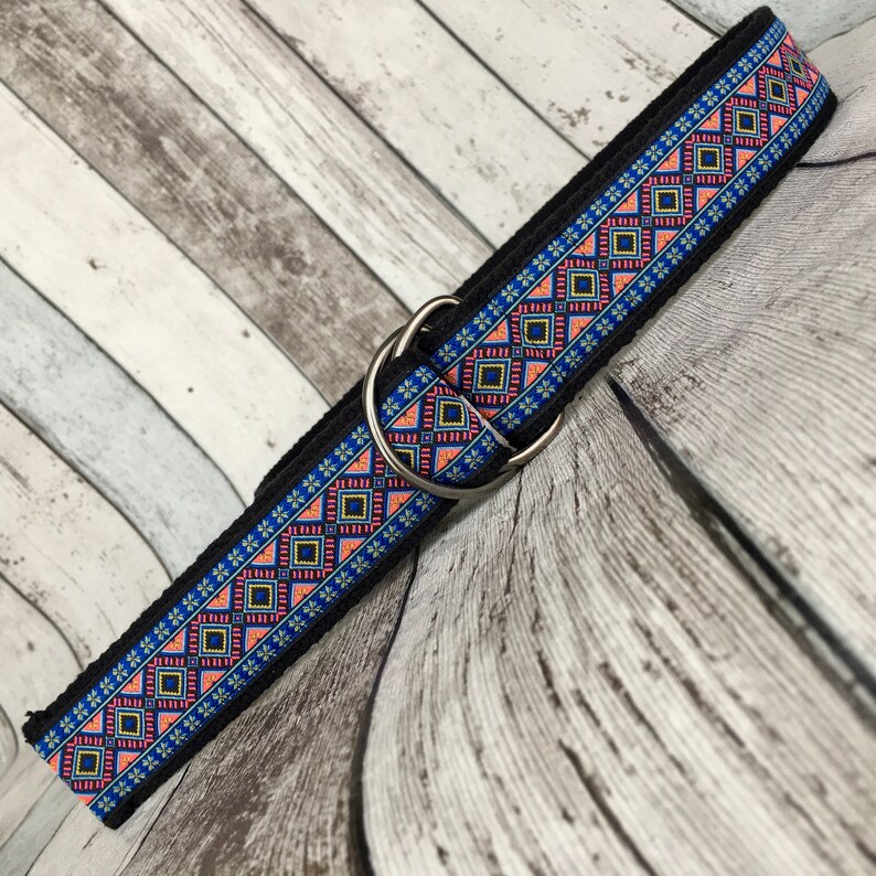 May include: A black woven belt with a colorful geometric pattern in shades of blue, pink, and yellow. The belt has a silver D-ring buckle.
