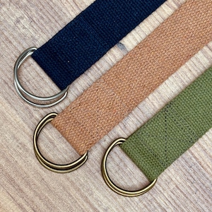 May include: Three canvas belts in navy blue, tan, and olive green, each with a metal D-ring closure. The belts are arranged diagonally against a light wood grain background, showcasing their texture and color variations.