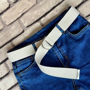 May include: A white canvas belt with a silver buckle on a pair of blue denim jeans.
