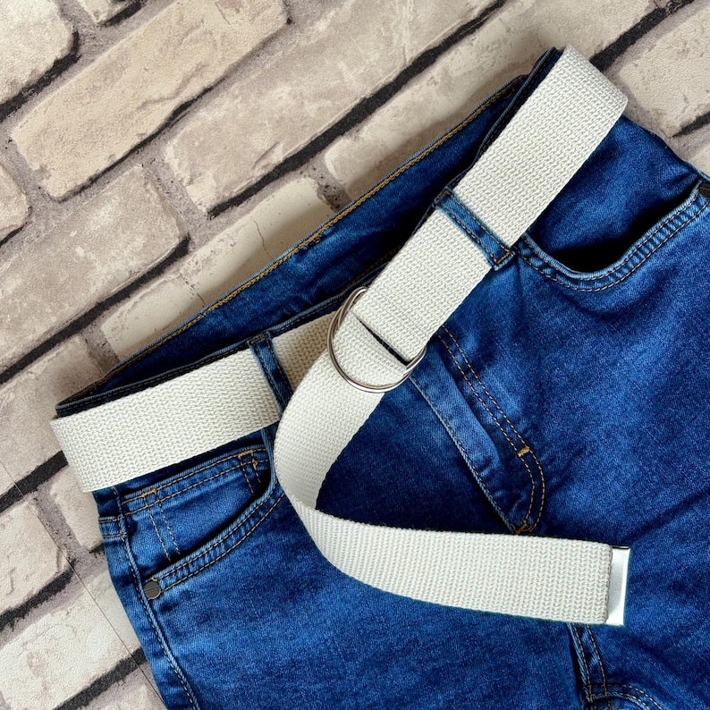 May include: A white canvas belt with a silver buckle on a pair of blue denim jeans.