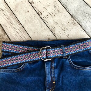 May include: A black woven belt with a pink, blue, and yellow geometric pattern. The belt is buckled and worn on a pair of blue jeans.