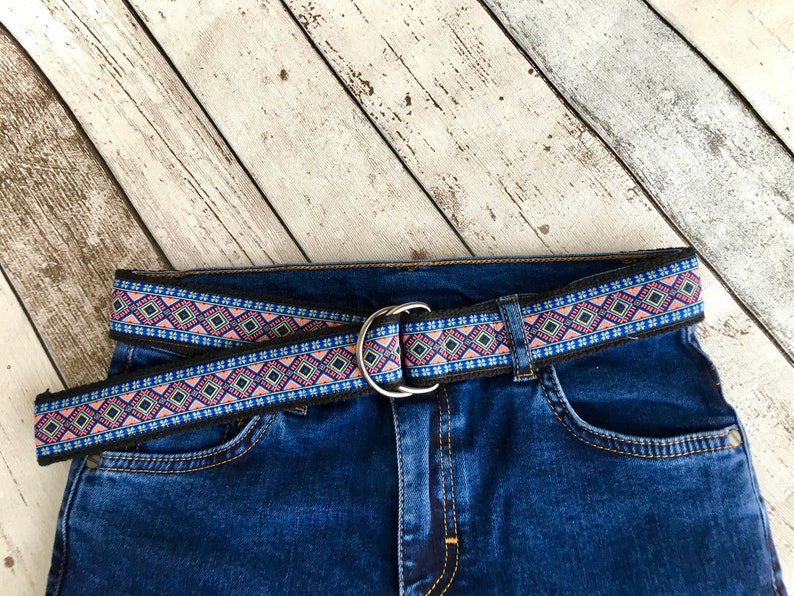 May include: A black woven belt with a pink, blue, and yellow geometric pattern. The belt is buckled and worn on a pair of blue jeans.