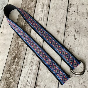 May include: A black woven guitar strap with a colorful geometric pattern in shades of pink, blue, and yellow. The strap has a silver metal D-ring.