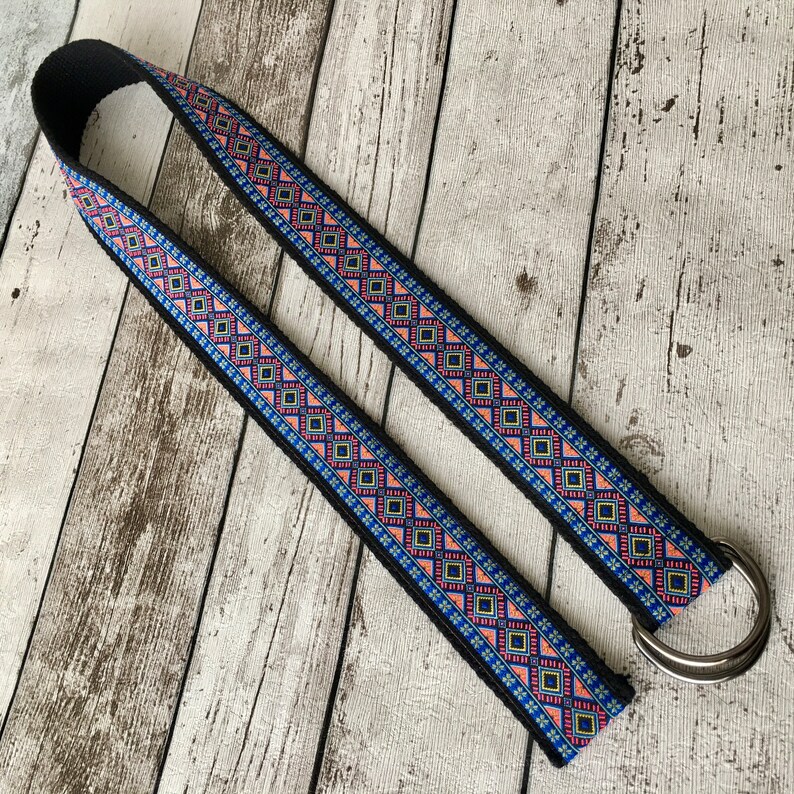 May include: A black woven guitar strap with a colorful geometric pattern in shades of pink, blue, and yellow. The strap has a silver metal D-ring.