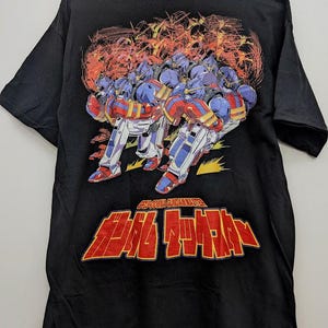 May include: A black short-sleeved t-shirt featuring a graphic print of multiple blue, red, and white robot figures in action. Below the robots, the text reads GF13-006NA GUNDAM MAXTER in white and Japanese characters in large red font with yellow outlines.