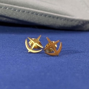 Puede incluir: Un par de pendientes dorados. Uno presenta un diseño de estrella y el otro un diseño de ancla. Los pendientes se muestran sobre una superficie azul.