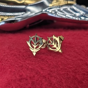 May include: Two gold-colored metal earrings with a stylized leaf design. The earrings are on a red surface. The earrings have a detailed, intricate design and a polished finish.