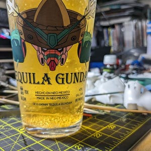 Puede incluir: Un vaso de vidrio transparente con un líquido amarillo en su interior. El vaso tiene un gráfico de un robot de dibujos animados con un sombrero. El texto en el vaso dice "Tequila Gundam" y "Hecho en Neo Mexico".