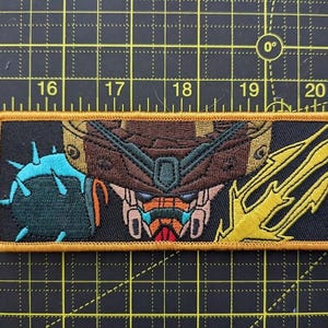 May include: Embroidered patch featuring a detailed robot face design with brown, orange, and teal accents. The patch has a black background and a gold border. The design includes a spiky blue object and a yellow lightning bolt.