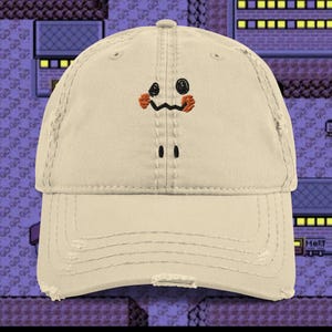 Op de afbeelding: Een beige baseballcap met een geborduurd Mimikyu-gezicht. Het ontwerp omvat zwarte ogen, een zwarte zigzagmond en oranje wangen. De cap heeft een gebogen klep en een versleten uitstraling, wat een vintage effect geeft.