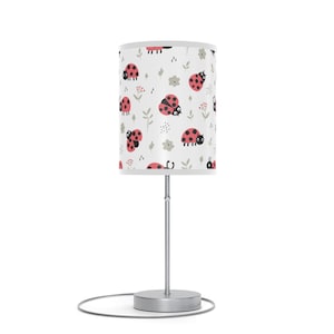 May include: A small table lamp with a white lampshade featuring a repeating pattern of red ladybugs, gray flowers, and green leaves. The lamp has a silver base and stem, and a round, silver base. The lamp is on a white background.