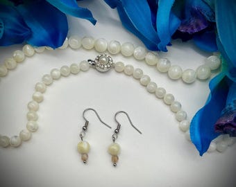 May include: A white mother of pearl necklace with a silver clasp and matching earrings with a single pearl drop. The necklace and earrings are displayed on a white surface with blue fabric in the background.