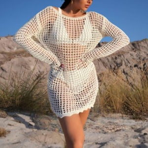 May include: A white crochet beach cover-up with long sleeves and a loose fit. The cover-up is made of a lightweight, breathable fabric and has a delicate, open-weave design. The model is wearing a white bikini top underneath the cover-up and a colorful striped hat.