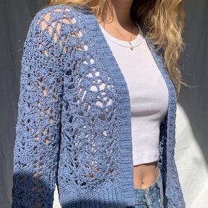 May include: A light blue crochet cardigan with an open weave design. The cardigan has long sleeves and a relaxed fit. The garment is worn over a white top and paired with blue jeans. The cardigan's color is a soft, muted blue.