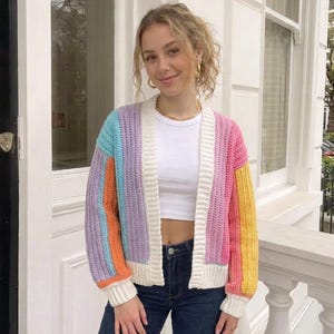 Puede incluir: Un c&aacute;rdigan de ganchillo de manga larga con rayas verticales en azul claro, naranja, lavanda, rosa y amarillo. La prenda tiene un borde acanalado blanco a lo largo de la abertura frontal, los pu&ntilde;os y el dobladillo, sobre un top corto blanco.