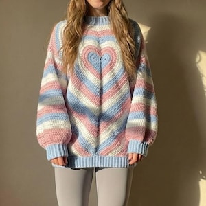 May include: A handmade crochet sweater with a heart design. The sweater features stripes of light blue, pink, and white. The sleeves are long and the fit is loose. The model is wearing grey leggings. The sweater is a cosy, oversized fit.