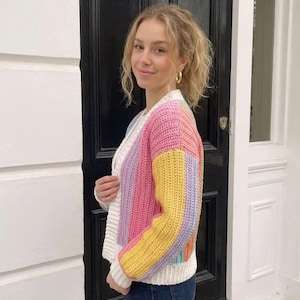 Puede incluir: Un c&aacute;rdigan de ganchillo de manga larga con rayas verticales en rosa pastel, amarillo, azul claro y lavanda. La prenda tiene un cuello, pu&ntilde;os y dobladillo acanalados en color blanco, con un cierre frontal de botones.