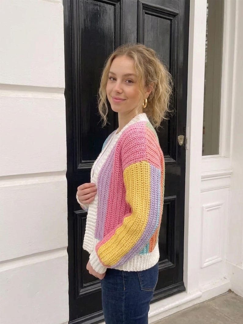 Puede incluir: Un c&aacute;rdigan de ganchillo de manga larga con rayas verticales en rosa pastel, amarillo, azul claro y lavanda. La prenda tiene un cuello, pu&ntilde;os y dobladillo acanalados en color blanco, con un cierre frontal de botones.