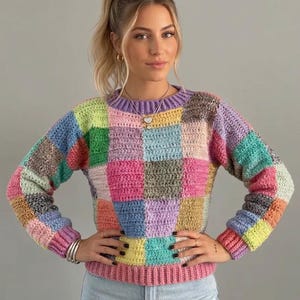 Op de afbeelding: Een gehaakte trui met lange mouwen en een veelkleurig patchworkontwerp met vierkanten in tinten roze, geel, blauw, groen, paars en grijs. Het kledingstuk heeft een geribbelde ronde hals, geribbelde boorden en een geribbelde roze zoom.