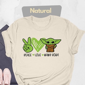 K&ouml;nnte beinhalten: Ein beiges T-Shirt mit einem gr&uuml;nen Peace-Zeichen, einem Herzen und einem Cartoon-Bild von Baby Yoda. Der Text auf dem Shirt lautet "Peace Love Baby Yoda".