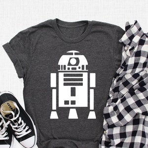 May include: A dark gray t-shirt with a white graphic of R2-D2, a robot from the Star Wars franchise. The shirt is laid flat on a gray surface with a black and white plaid shirt and black sneakers.